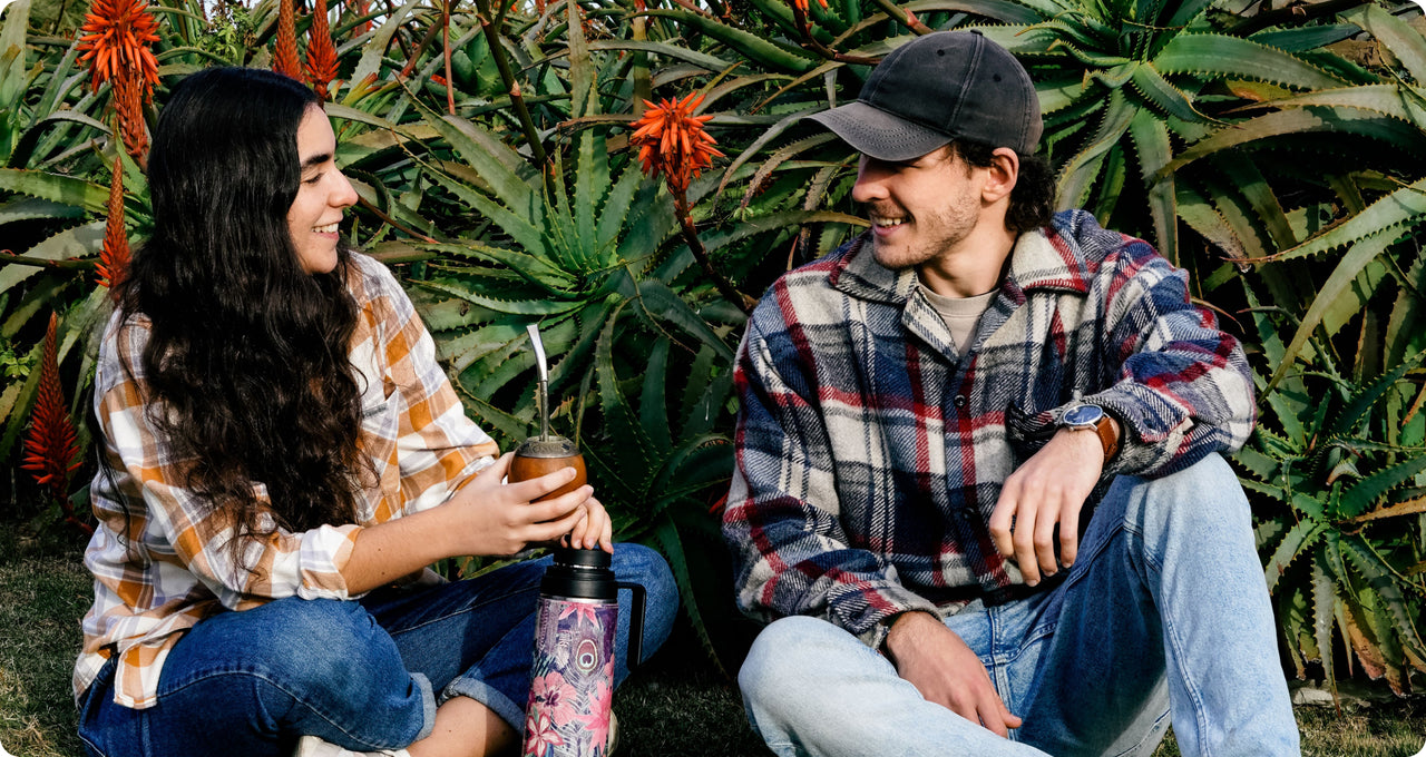 A young woman and man sit on the grass, smiling at each other, surrounded by lush green aloe plants with vibrant orange flowers. The woman holds a traditional yerba mate gourd with a metal straw, and a thermos with a colorful design sits beside her. They are both dressed in plaid shirts and jeans, creating a cozy and relaxed outdoor setting.