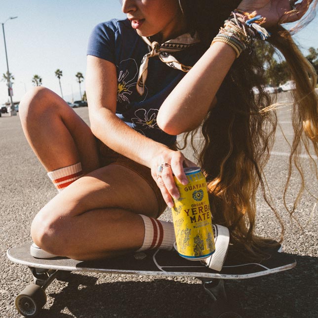 A young woman with long, wavy hair sits on a skateboard, holding a can of Guayakà Yerba Mate. She is wearing retro-style socks and bracelets, with palm trees and a sunny outdoor setting in the background.
