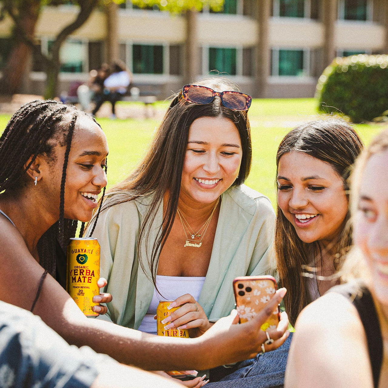 Four people sit on the grass, smiling as they look at a phone. Two hold high-energy Guayakí Yerba Mate cans, enjoying the sunny day outdoors with a building in the background. The scene exudes a lively vibe, thanks to their refreshing organic products.