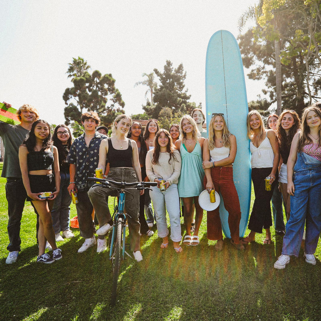 A group of young adults and teenagers gather in a sunny park holding Guayaki. They are smiling and standing together, with one person holding a bike and another holding a surfboard. Trees and grass are visible in the background.