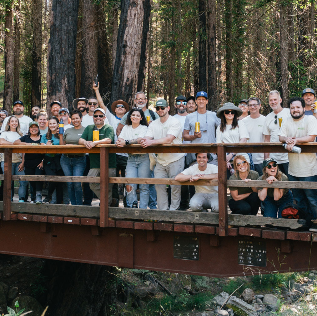 A large group of people stand together on a wooden bridge in a forest, smiling and holding cans of Guayakà Yerba Mate. They are dressed casually, with some wearing hats and sunglasses, surrounded by tall trees.