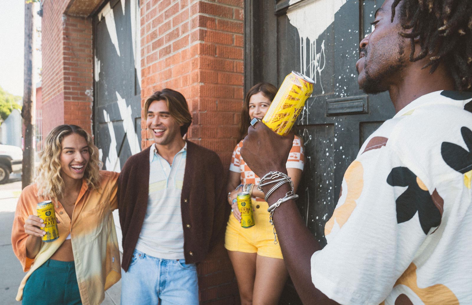 4 people standing next 2 a building laughing and talking. They are holding and drinking Guayaki cans.