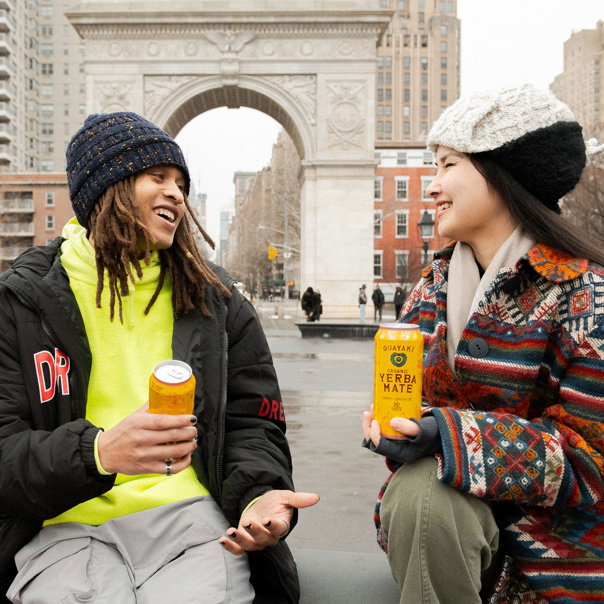 2 people smiling a talking in the city holding Guayaki Yerba Mate cans.