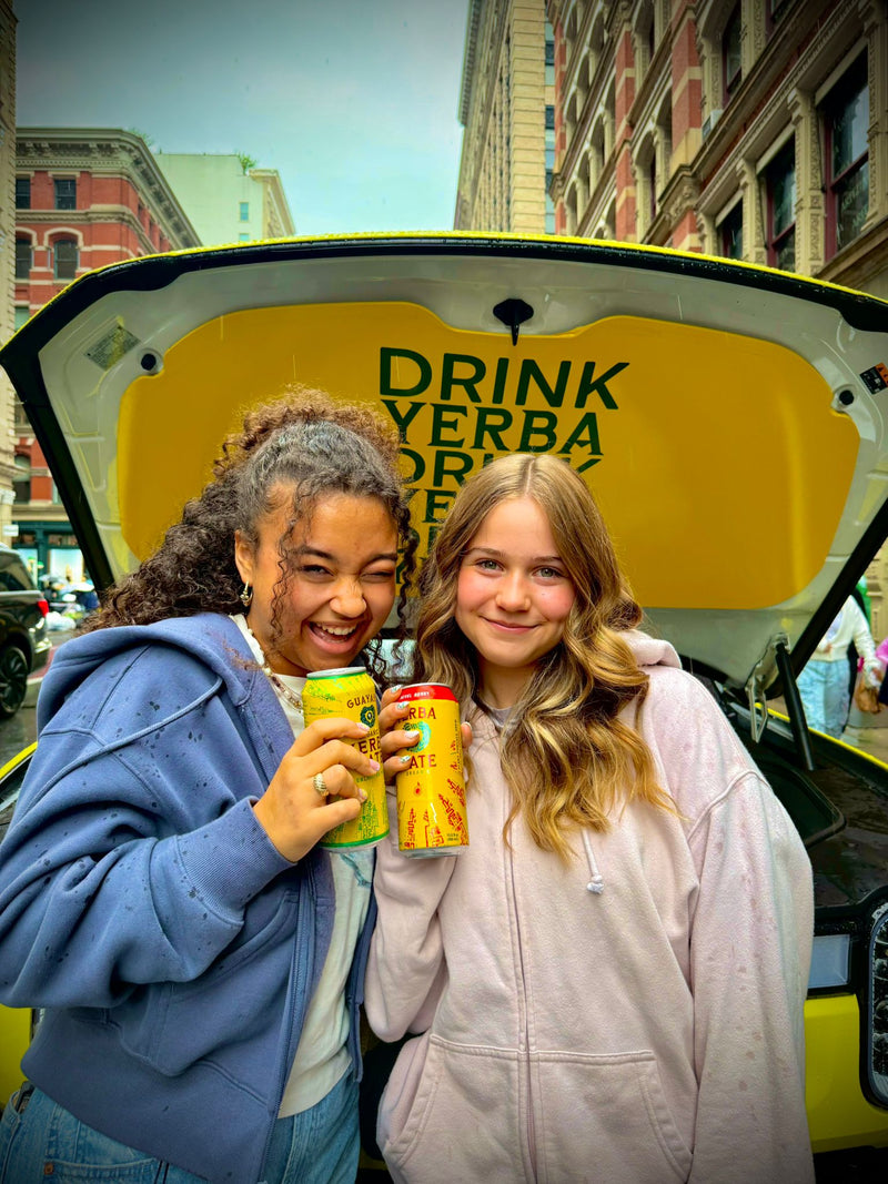 Two people holding drinks in front of a yellow vehicle with 'Drink Yerba' text.