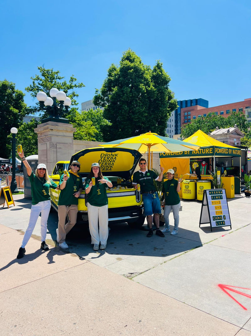 Group of people in green shirts and white pants posing in front of a yellow food truck on a sunny day.
