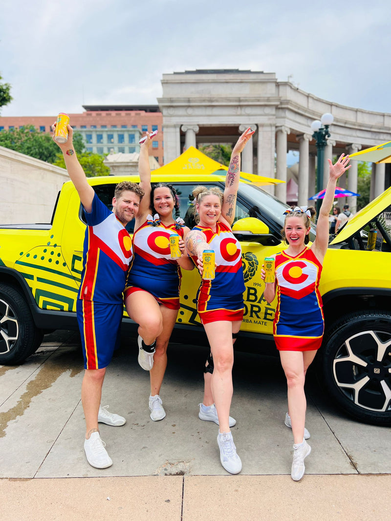 Four people in colorful outfits with circular designs are posing in front of a yellow vehicle.