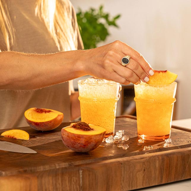 A woman garnishes two iced yerba mate drinks with fresh peach slices on a wooden cutting board. Halved peaches and ice cubes are scattered around, with a warm and inviting kitchen setting in the background.