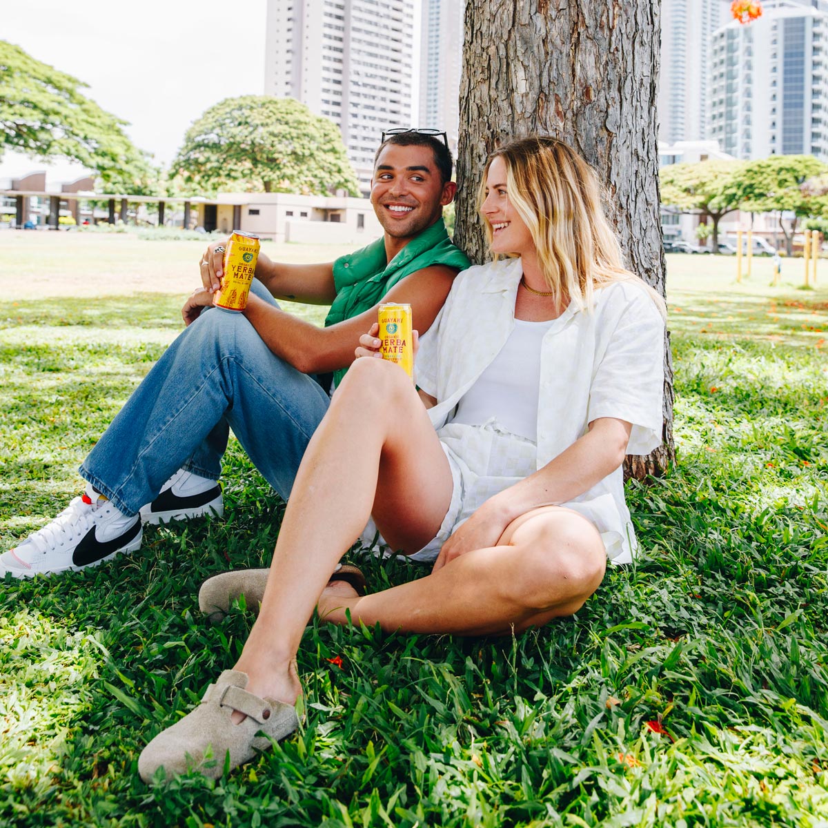 2 people sitting in grass and leaning on a tree holding Guayaki Yerb Mate