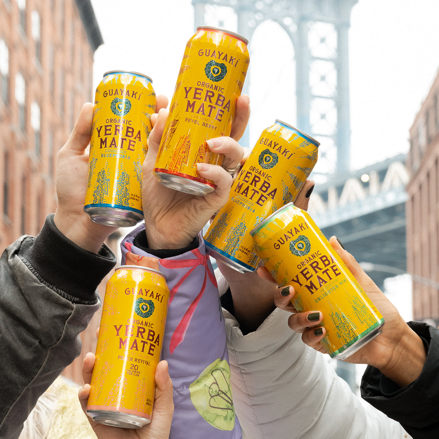 A group of hands raises cans of Yerba Madre in a celebratory toast, with the a bridge framed between brick buildings in the background.