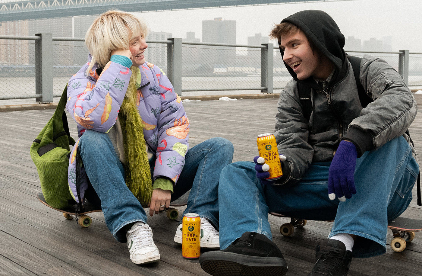 Two friends sit on skateboards on a wooden pier, smiling and chatting while holding cans of Yerba Madre. A bridge and city skyline appear in the misty background.